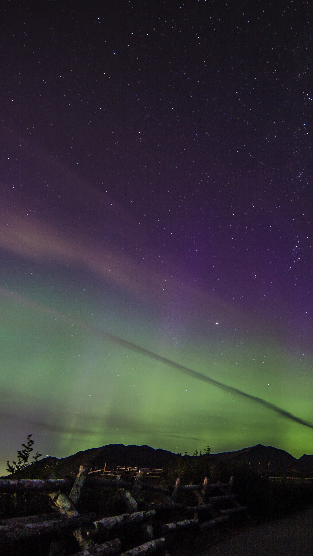 A night landscape photograph showing the aurora borealis with green, purple, and pink light over dark mountains and a starry sky, with a rustic wooden fence in the foreground.