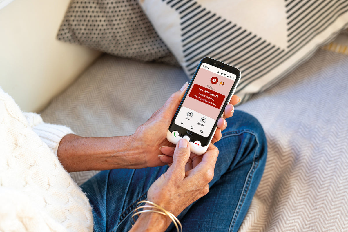 A person sitting on a couch holds a smartphone showing a red and white app interface. The person is wearing a white knit sweater and blue jeans, with patterned pillows and a textured blanket in the background.